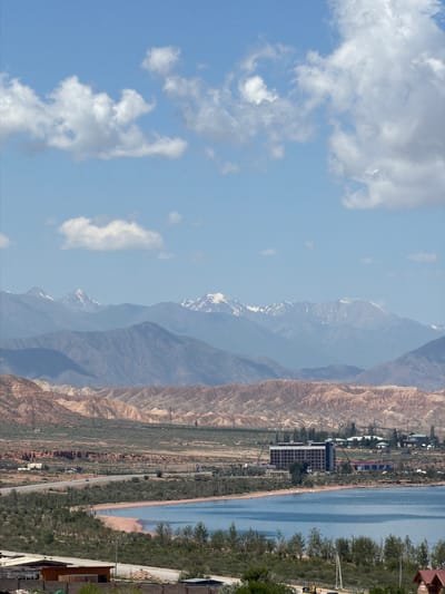 The image depicts a scenic view of Kaji-Say, Kyrgyzstan, with a clear blue sky dotted with white clouds. In the foreground, a tranquil body of water, likely Issyk-Kul Lake, is visible, with a sandy shoreline fringed by greenery. Beyond the lake, a road winds through a landscape of rolling hills and arid terrain. A prominent modern building, possibly a hotel or resort, stands near the shoreline, surrounded by other structures and vegetation. In the background, majestic snow-capped mountains rise against the horizon, their rugged peaks forming a dramatic backdrop to the scene. The overall impression is one of natural beauty and developing infrastructure in a mountainous region.