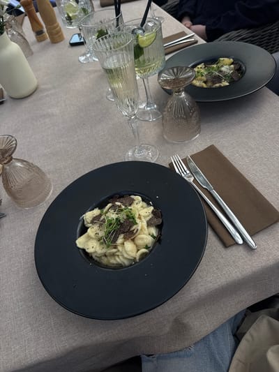 The scene depicts a dining table arrangement, likely in a restaurant, featuring pasta dishes topped with what appears to be truffle shavings. The location is Konstancin-Jeziorna, Poland, as indicated in metadata not provided but which can be deduced by the image set name.
On the table, there are two dark-colored plates, each containing a serving of pasta. Silverware (fork and knife) rests on a brown napkin beside one of the plates. Several glasses are visible, some filled with a clear liquid (possibly a cocktail) containing lime slices. Also visible are a bottle and other tableware items. The tabletop has a light, textured fabric covering. In the background, a person wearing jeans and a beige jacket is partially visible. Another person in a dark blue garment is seated on the left.