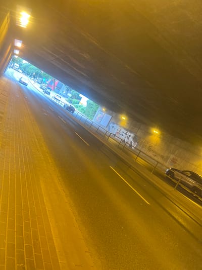 The image depicts a view from within a tunnel on a multi-lane road in Herne, Germany. The tunnel has a concrete ceiling and walls, illuminated by artificial lighting. The road surface is asphalt, with visible lane markings. On the left side of the road, there is a pedestrian walkway paved with yellow bricks. Several vehicles are present on the road, including cars and a truck in the distance. The tunnel walls are adorned with graffiti. The overall scene suggests a typical urban environment with traffic flow through a tunnel.