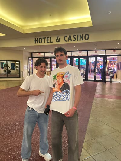 Two men are standing in front of the entrance to the Seminole Hard Rock Hotel & Casino in Hollywood, Florida. One man is wearing a white t-shirt with an image on it and grey pants. The other man is wearing a plain white t-shirt and jeans. The entrance to the casino is visible in the background.