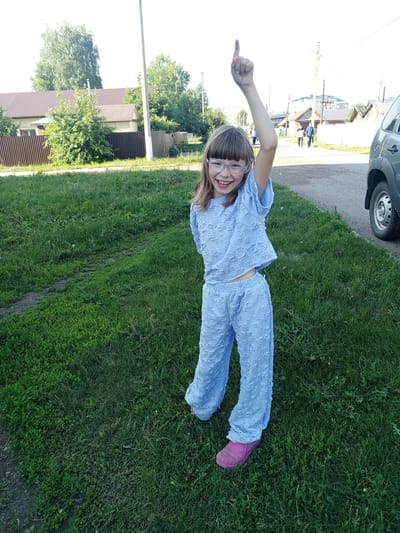 A young girl, wearing glasses and a light blue textured two-piece outfit consisting of a short-sleeved top and pants, stands on a patch of green grass. Her right arm is raised with her index finger pointing upwards. She is smiling. The background shows a street with a few buildings and trees, and a gray SUV parked on the right side of the image. The location is the city of Dyurtyuli, Russia.