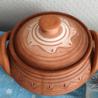 A close-up, high-angle shot shows a terra cotta pot with a lid. The pot has two handles and decorative concentric rings and wave-like patterns with small oval indentations. The lid has a rounded knob and similar decorative rings. The pot sits on a blue cloth with a snowflake pattern. The background is a white textured surface. This image was taken in Tohatin, Moldova.