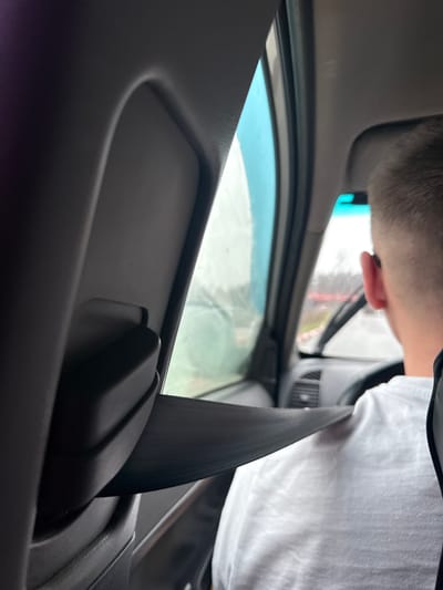 This image captures the interior of a vehicle, likely a car or van, viewed from a low angle, possibly from a rear seat looking towards the front passenger side. In the foreground, a prominent black seatbelt stretches diagonally across the lower right. A person, seen from the back of their head with short hair and a visible ear, occupies the upper part of the frame, wearing a white t-shirt.
The person appears to be positioned near the vehicle's front side, possibly leaning forward or looking out the side window. The car's grey interior door panel and window frame are visible, with the window showing a blurred view of the outside. The exterior scene is indistinct, suggesting either motion or overcast weather conditions during the daytime. A light blue tint, possibly from a mirror or part of the vehicle's structure, is visible near the person's head. No specific activity beyond observing the outside is clearly discernible. There are no visible texts in the image.