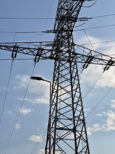 The image depicts a steel lattice electricity pylon silhouetted against a blue sky with scattered clouds. The pylon, presumably located in Bettembourg, Luxembourg, judging from the location information, supports high-voltage power lines. A street light is mounted on the pylon. The pylon structure consists of interconnected steel beams and cross braces. Insulators are attached to the tower to isolate the wires from the structure. Overhead power lines extend horizontally across the frame.