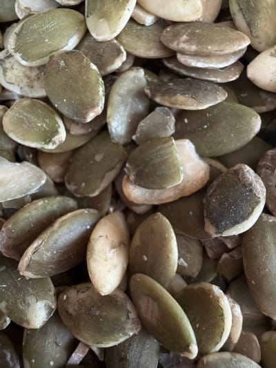 A close-up, overhead shot of raw, hulled pumpkin seeds fills the frame. The seeds are an oval shape with a slightly flattened, curved surface. Their color ranges from a pale off-white to a muted olive green, with some seeds exhibiting brown or greyish tinges, likely from natural variation or slight imperfections in the hulling process. The texture appears smooth, with some seeds showing subtle striations or a slightly grainy surface. The lighting casts soft shadows, highlighting the rounded forms of the seeds and creating a sense of depth within the pile. There are no people or specific environmental features visible, indicating the image is a still life focused solely on the produce. The location in Капцёўскі сельскі Савет, Belarus, is not visually represented in this image.