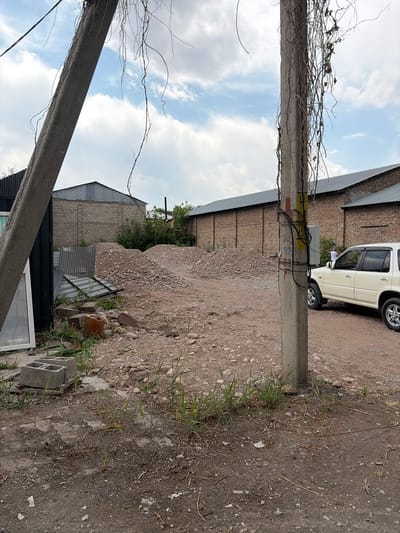The image depicts an outdoor scene in the city of Kant, Kyrgyzstan, characterized by a construction or demolition site. In the foreground, a concrete utility pole stands prominently, adorned with numerous wires and some dried, trailing vines. To the left, a large, angled concrete pillar is visible, along with parts of a dark, rectangular structure that appears to be a building or shed with a large window. The ground is a mixture of dirt, gravel, and scattered debris, including concrete blocks, bricks, and metal sheeting.
In the mid-ground, large mounds of rubble and small stones dominate the landscape, suggesting excavation or demolition activities. Behind the rubble, several long, low-rise brick buildings with corrugated metal roofs stretch across the scene. A white SUV is parked on the dirt road to the right of the utility pole, facing left. A person in a white shirt can be seen near the rear of the SUV, interacting with an electrical box. The sky above is partially cloudy, with a mix of blue sky and white clouds. The overall impression is one of an industrial or construction area in a state of disarray.