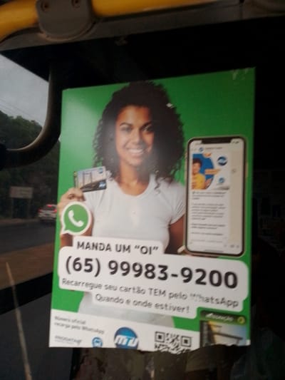 A woman with dark, curly hair and a white t-shirt is holding a mobile phone. She is smiling and looking at the viewer. On the phone screen, there is a WhatsApp chat. The woman is holding what appears to be a credit card or a transit card in her left hand. The advertisement is green and white. A phone number, "(65) 99983-9200", is displayed prominently on the ad. Below the phone number, it says "Recarregue seu cartão TEM pelo WhatsApp. Quando e onde estiver!". There is also a QR code in the bottom right corner. The advertisement is likely placed on public transportation, possibly a bus, as indicated by the yellow handle at the top of the image. The background shows a slightly blurred view of a street with cars and buildings, suggesting an urban environment. The context of the phone number format and the brand "TEM" strongly indicates the location is Cuiabá, Brazil, where the "Cartão TEM" (Transporte em Metrô) is a common public transport card.
