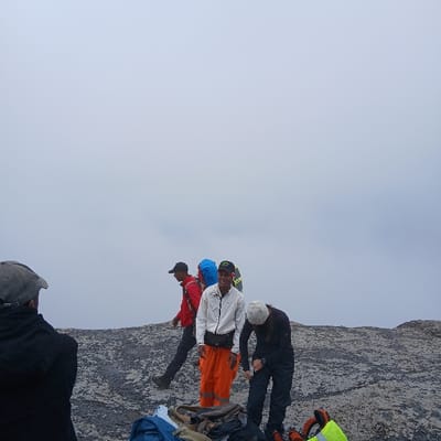 The image depicts a group of five individuals at Gilman's Point, Tanzania, under an overcast sky that obscures the horizon. The ground is rocky and dark grey, indicative of high-altitude terrain.
In the foreground, with their back to the camera, is an individual wearing a dark jacket and a grey baseball cap. To their right, in the mid-ground, a person wearing a red jacket, dark pants, and a dark baseball cap is walking from left to right, carrying a blue backpack with a bright yellow accent. Adjacent to them, another individual stands facing the camera, smiling, dressed in a white jacket, bright orange pants with reflective strips, and a dark baseball cap. A black fanny pack is visible around their waist, and a yellow/green backpack is behind them. To the right of this smiling individual, another person with long hair or a grey cap, dressed in dark clothing, is bent over, appearing to adjust something on their leg or boot with a white object in their hand. Various items of gear, including a bright yellow/green bag or vest, are laid out on the rocky ground in the foreground and mid-ground. The overall scene suggests a high-altitude expedition or trek.