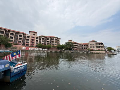 A scenic view of Sungai Melaka featuring calm water reflecting the sky and surrounding architecture. Buildings with red-tiled roofs line the riverbank. A boat with the Malaysian flag and "Hatten Hotel" written on its side is partially visible in the foreground. A sign reading "#i@sungaimelaka" adds to the tourist-friendly atmosphere.