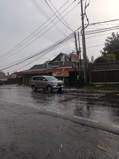The image depicts a street scene in Cugenang, Indonesia, during rainfall. The asphalt road surface is thoroughly wet, reflecting the overcast sky and ambient light. White dashed lane markings are visible on the road, with water splashes evident, particularly around a moving vehicle. A silver/grey multi-purpose vehicle (MPV), resembling a Toyota Innova, is traveling from left to right in the midground, its front wheels creating visible water spray.
On the right side of the road, several buildings are visible. The most prominent is a two-story structure with a brown tiled roof and a light-colored facade. Below this, a commercial establishment features a red and white banner displaying food items and text, including "AYAM GEPREK" (smashed fried chicken). A separate red sign, partially obscured, reads "AYAM NAGA" (Dragon Chicken) with a phone number. A tall, dark utility pole stands directly in front of these buildings, heavily laden with a dense network of black electrical and communication wires that crisscross the sky. A red conduit is also attached to the pole. Further to the right, a concrete fence with decorative block patterns separates the road from lush green foliage and trees. The sky is uniformly overcast and light grey, indicating an ongoing or recent rain event. No human figures are present in the scene.
