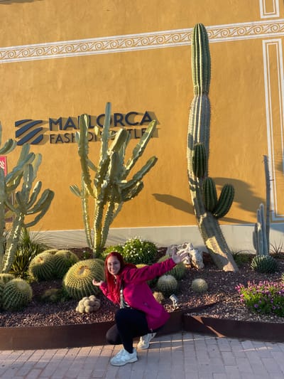 An outdoor scene at the Mallorca Fashion Outlet in Marratxí, Spain, depicts a single female subject posing in a landscaped area. The subject, a young adult with reddish-orange hair and glasses, is positioned in a crouch facing the camera, smiling with both arms extended outward and palms open. She wears a dark pink long-sleeved zip-up jacket, black leggings, and white athletic shoes.
Behind her, a substantial cactus garden occupies a raised planter with dark metal edging. This garden features numerous large cacti, including tall columnar varieties, multi-branched specimens, and several prominent spherical barrel cacti. Smaller cacti, agave-like plants, and patches of white-flowering and purple-flowering shrubs are interspersed amongst dark reddish-brown gravel ground cover and light-colored rocks.
The background consists of a building facade painted in a warm, muted yellow or orange. Mounted on this facade are the dark, raised letters spelling "MALLORCA FASHION OUTLET" with a blue 'Z' shaped logo preceding "MALLORCA." A horizontal decorative band with a white scroll pattern runs across the upper portion of the wall. Strong, elongated shadows from the cacti and the subject are cast across the ground and up the building, indicating bright sunlight, likely late afternoon or early morning. The immediate foreground shows a paved walkway composed of light-colored rectangular pavers.