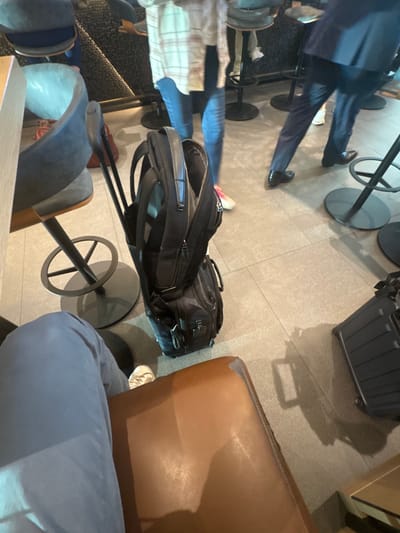 The image depicts an indoor scene within a commercial establishment, identified as being in Arlington, United States. The perspective is from a seated individual whose light blue trousers and white athletic shoes are visible in the lower left, occupying a brown leather-upholstered chair.
Centered on the light gray tiled floor is a black rolling luggage bag with a matching black backpack placed on top, from which a single black-handled golf club protrudes. To the right, a dark, ribbed hard-shell rolling suitcase is partially visible. A light gray upholstered bar stool with a black metal base is situated to the left, with a small red bag resting on the floor nearby.
In the mid-ground, several individuals are present. One person wears blue jeans, a light plaid shirt, and pink athletic shoes. Another, partially blurred due to movement, is dressed in dark trousers, a dark jacket, and black dress shoes, appearing to be walking. Other individuals' legs are visible in the background, seated on black metal bar stools against a dark, textured wall. The overall environment suggests a lounge, bar, or airport waiting area.