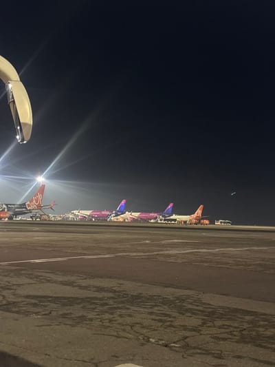 An outdoor scene depicts an airport tarmac at night, situated in Bubuieci, Moldova. The image is captured from a slightly elevated vantage point, with a light-colored, curved object, likely a part of the originating aircraft or airport infrastructure, partially present in the upper-left foreground. Intense artificial lighting illuminates the area, generating significant lens flares. The paved tarmac surface in the foreground exhibits visible cracks and linear markings.

In the midground, a line of at least five parked passenger aircraft is observed. From left to right: a dark grey aircraft with an orange tail and "SkyUp" branding visible on its fuselage. A mobile ground stair is connected to this aircraft. Following it are three distinct magenta and white aircraft, characteristic of Wizz Air livery. To their right, an aircraft with a predominantly white fuselage and an orange tail is visible. Various ground support equipment, including small utility vehicles and a white bus positioned on the far right, are arrayed near the aircraft, indicating ongoing or paused ground operations. No identifiable individuals are distinctly visible within the frame.