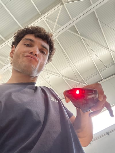 The image depicts an adult male taking a selfie within an industrial setting in Polígon Industrial De Son Castello, Spain. The male, visible from the chest up, has curly brown hair and light skin, and is making a pouting facial expression directly towards the camera. He is wearing a dark blue t-shirt with white lettering on the left chest. His right hand holds a black, angular electronic device with a prominent, illuminated red light on its front face, positioned in the lower right portion of the frame. The background features a high, light-colored ceiling composed of a metal truss structure and corrugated panels, characteristic of a warehouse or large industrial building.