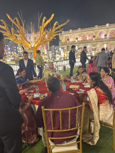 People are seated at banquet tables enjoying a meal at an outdoor event at night. The tables are covered in red cloths and adorned with water bottles, plates, and what appear to be decorative accessories. Individuals wear traditional Indian attire. A building with illuminated arches and floral arrangements forms the backdrop. An elaborate gold-colored structure is on the left, with its branches reaching towards the sky. Other event attendees stand or walk around in the background.
