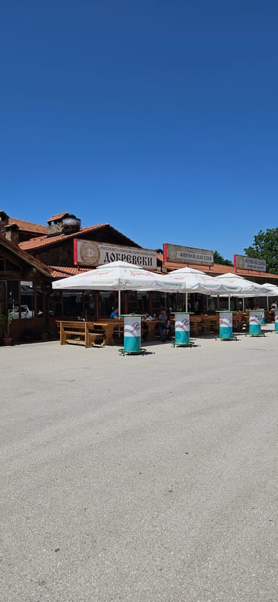 In Zlatna Panega, Bulgaria, this image depicts the exterior of a restaurant and shops under a clear blue sky. The building features a traditional tiled roof and stone construction. In the foreground, outdoor seating is provided with tables and benches under large white umbrellas, some of which have logos. Several cylindrical trash receptacles with teal and white designs are placed around the seating area. A few individuals are seated at the tables, appearing to be customers. Signs above the restaurant entrance are visible, indicating "ДОБРЕВСКИ" (DOBREVSKI) and other text in Bulgarian, identifying it as a restaurant and a shop. The overall scene suggests a commercial establishment with an outdoor dining area.