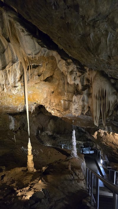 This image depicts the interior of a cave, likely a tourist attraction given the presence of a metal railing and stairs in the background. The cave walls are rough and textured, with formations of stalactites and stalagmites visible throughout. In the foreground, a prominent stalactite hangs horizontally, illuminated by a light source that casts a distinct glow. Several smaller stalactites are also visible, appearing as white, needle-like structures on the cave walls and ceiling. The lighting is artificial, designed to highlight the cave's natural features. The overall scene conveys a sense of awe and natural wonder. The location is identified as Vavřinec, Czechia, which likely refers to a specific cave system in that region. There is no visible text in the image.