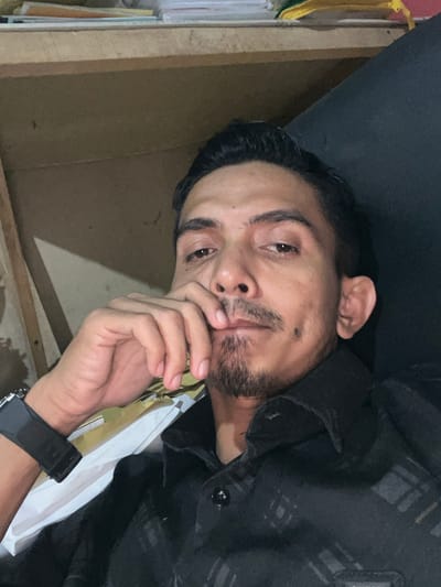 A close-up, waist-up, selfie-style indoor photograph depicts an adult male in Alueduamuka S, Indonesia. The subject, positioned centrally, is observed with dark hair, dark eyes, and facial hair consisting of a mustache and a goatee. He is wearing a dark, possibly black, collared shirt with a subtle checkered or patterned design. A black digital watch is visible on his left wrist. His left hand is raised, with the index and middle fingertips touching his upper lip and chin area. He looks directly forward towards the camera. The background to the left appears to be a light-colored wooden or laminate surface, possibly shelving, with several stacks of papers or books visible on an upper ledge. The background to the right is dark and indistinct, suggesting a wall or furniture.
