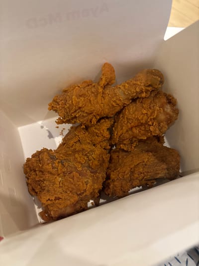 The image shows four pieces of fried chicken in a white, open cardboard container. The chicken pieces are golden brown and appear crispy. The container is positioned at an angle, showing its interior and some text partially visible on one side. The background shows the edges of what looks like table. No people are visible. The location cannot be determined.