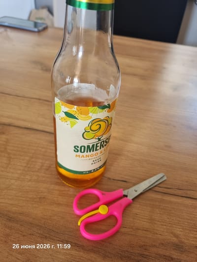 A close-up, high-angle shot shows a glass bottle of Somersby Mango & Lime beer on a wooden table. The bottle is partially filled with amber liquid and has a white label with colorful fruit illustrations. To the right of the bottle are a pair of pink children's scissors with yellow accents. The background is blurred, showing a dark surface and a light-colored object. A timestamp in the lower left corner reads "26 июня 2026 г. 11:59". The location is Radzymin, Poland.