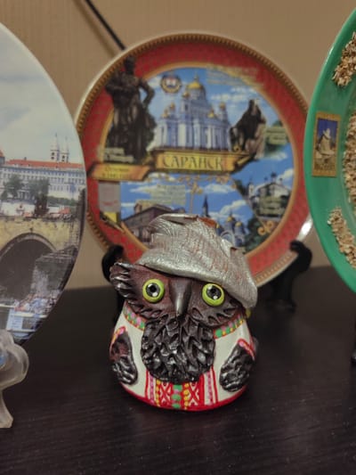 A small, owl-shaped figurine is positioned in the foreground, facing the viewer. The figurine has large green eyes with black pupils and wears a silver-colored hat and a white tunic adorned with red and green embroidered patterns. Its body is dark brown with textured wings. The figurine is placed on a dark, flat surface.
Behind the figurine, a circular decorative plate is visible. The plate features a central image of a blue sky with white clouds, a gold-domed building, and a dark statue. Text in Russian, "Саранск," is written on the plate. The plate has a red border with a gold decorative pattern.
To the left of this plate, another decorative plate is partially visible. It is oval-shaped and depicts a bridge over a body of water, with buildings in the background.
To the right of the central plate, a green decorative plate is visible, filled with what appears to be small grains or seeds. A small framed image of a person in a hat is attached to this green plate.
The background consists of a plain, light-colored wall. The lighting suggests an indoor setting. The overall scene appears to be a display of decorative plates and a figurine on a shelf. The specific location in the city of Ostafievo, Russia, is not discernible from the visual information provided in the image.