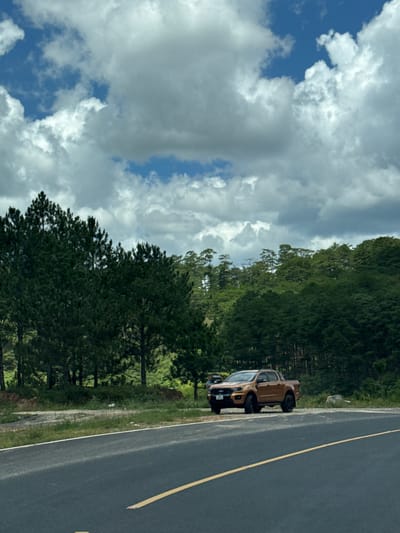 The image depicts a brown Ford Ranger pickup truck parked on the shoulder of a paved road. The truck is positioned on the right side of the frame, facing left. The road has a yellow lane divider. In the background, a dense forest with tall pine trees extends up a hillside. The sky is partly cloudy with large, white cumulus clouds against a blue backdrop. The location appears to be a rural or mountainous area, possibly on the outskirts of Đa Bla, Vietnam, given the presence of pine trees common in the Central Highlands region. There are no people clearly visible in the immediate vicinity of the truck. The overall scene suggests a stop along a scenic route.