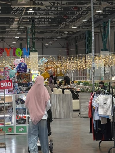 The image depicts an indoor scene, likely a market or store, with numerous people present. The setting appears to be a large, open space with a high ceiling, characterized by industrial-style metal shelving and overhead lighting. Strands of small, yellow lights are strung across the space, creating a festive or decorative atmosphere.
Several individuals are visible, some standing, others seated. One person in the foreground is wearing a pink hooded garment and blue jeans, pushing a trolley. Other people are browsing or interacting in the background, where tables and chairs suggest a seating area. The overall impression is one of a busy commercial environment.
Objects in the scene include clothing displayed on racks, various products on shelves, and decorative items like balloons and the string lights. Some of the products appear to be toys or children's items, judging by the colorful packaging and characters visible. There is also a red sign with white text that reads "500". The time of day is not explicitly indicated, but the indoor lighting suggests it could be day or evening. No specific weather conditions can be inferred from the image. The provided location context is Kant, Kyrgyzstan.