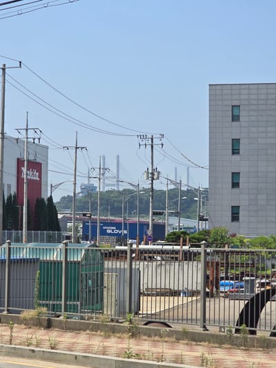 A wide outdoor shot shows a street-level view of a commercial or industrial area on a clear day. In the foreground, a gray metal fence runs horizontally across the image, with a concrete planter box containing sparse vegetation and a sidewalk of red and beige patterned pavers behind it. Beyond the fence, a green-tinted bus stop shelter with a curved roof is visible, as are other industrial elements like metal scaffolding and a portion of a blue shipping container with "HYUNDAI GLOVIS" printed on it. To the left, a large white building with a prominent red "Makita" logo stands, flanked by evergreen trees. A network of electrical poles and wires crisscrosses the upper portion of the scene. In the background, a large, modern gray building with multiple windows is visible on the right side of the frame. Further in the distance, lush green hills rise, and a light blue sky dominates the upper half of the image. A suspension bridge with tall towers can be seen in the far background, suggesting proximity to a major waterway or harbor, consistent with the industrial landscape of Pyeongtaek-si, South Korea. A black object, possibly a dog or a large tarp, is partially visible in the lower right corner.