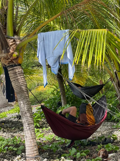 An overhead shot captures a person relaxing in a maroon hammock strung between palm trees on a beach. The individual, wearing black shorts and a brown and orange shirt, is partially visible, with their legs and feet propped up. Another black hammock hangs above, empty. Clothes are draped on a nearby palm tree, and a pair of black sandals rests on the rocky, vegetated ground beside the hammock. The scene is set in Babahie, Indonesia, characterized by lush palm trees and a tropical environment.