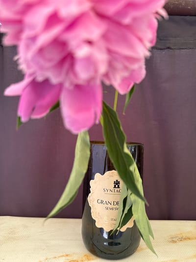 A close-up vertical shot displays a vibrant, out-of-focus pink peony occupying the upper portion of the frame, with its numerous ruffled petals obscuring much of the background. Green stems and leaves descend from the flower, partially draping over a dark-colored glass bottle positioned centrally in the lower half of the image. The bottle features a light-colored, ornate label with a black castle silhouette at the top, followed by the text "SYNTAC," "GRAN DE," "SEMI SV," and partially visible "ITAL." The background consists of a solid, dark reddish-purple wall or surface. The foreground is a light-colored, textured surface, possibly fabric, exhibiting several reddish-brown stains, predominantly on the right side. The overall depth of field is shallow, with the flower being the most blurred element.