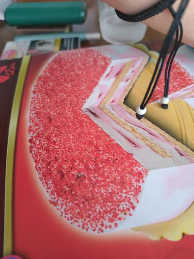 The image shows a close-up, angled view of a box of ice cream cake. The cake is depicted with layers of pink and yellow ice cream, covered in red sprinkles. The box is predominantly red with some yellow and green accents. In the background, a person's arm is visible, wearing a black braided bracelet, with black cords with white beads hanging down. The scene appears to be indoors, possibly on a table, with some blurred objects in the background, including what looks like a green cylindrical object and a glass. There is no information or visual cues to suggest the location in the city of Yagoda, Bulgaria.