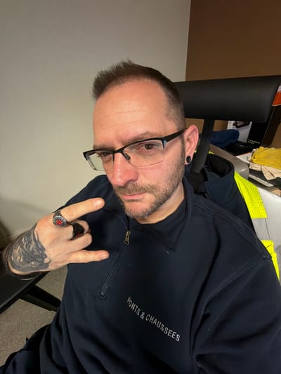 The image depicts a male individual, observed from the chest up, in an indoor environment located in Grevenmacher, Luxembourg. The subject has short, dark hair styled with a slight pompadour, a trimmed beard and mustache, and wears black-framed, semi-rimless glasses. Black plugs are present in both earlobes. He is wearing a dark blue quarter-zip pullover with the text "PONTS & CHAUSSEES" embroidered in white on the left chest. A yellow high-visibility safety vest with reflective strips is visible behind and over his right shoulder. The individual's left hand is raised, forming a "rock on" gesture with the index and pinky fingers extended. Two rings are visible on this hand: a black ring with a red circular emblem on the ring finger, and a black ring on the middle finger. Black and grey tattoo work is visible on the subject's left forearm/wrist. The background features a plain light-colored wall to the left and a brown wall or partition to the right. A black office chair headrest is visible behind the subject. Various items, including a blue cloth, a yellow cloth, and a partially visible white box with black text indicating "1.0hp Battery Charger," are present on a surface in the right background.