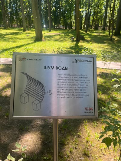 An outdoor informational sign about the "Sound of Water" is depicted in a park in Guryevsk, Russia. The sign is made of metal and mounted on a pole. It features text in Russian, an illustration of a water feature resembling a wave or cascade, and logos for "KURGAN-BALT" and "PROStal". The text discusses how the sounds of nature, particularly water, are relaxing and captivating, and encourages visitors to listen to the sound of a nearby stream to escape everyday worries and immerse themselves in a world of peace and harmony. Below the main text, a phone number "777-245" and a website "kurgan-balt.ru" are visible. The sign is surrounded by lush green grass and trees, suggesting a sunny day in a well-maintained park.