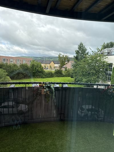 This is a vertical, outdoor photograph taken from a covered patio, looking out over a residential neighborhood. The foreground is dominated by the dark wooden beams and ceiling of the patio, with some bamboo or slatted screening visible on the right. Beyond the patio is a section of artificial grass.
The middle ground features a neatly mown green lawn, with several trees and shrubs. A dark, slatted fence runs along the edge of the lawn, with some decorative lights strung along it. A potted plant with green foliage hangs on the fence.
In the background, there are several residential buildings. One is a modern, cream-colored house with multiple windows, partially obscured by trees. Further to the left, there is a brick apartment building with many windows, and behind that, other houses with pitched roofs, indicative of a suburban or semi-rural setting. The landscape beyond the houses appears to be hilly, with fields and more scattered buildings visible in the distance.
The sky is overcast and appears to be a light gray, suggesting cloudy weather. The time of day is indeterminate due to the cloud cover, but it does not appear to be dawn or dusk. There is no text visible in the image. The overall impression is a somewhat muted, overcast day in a residential area.