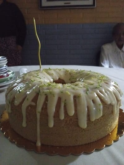 The image presents a ring cake on a golden scalloped plate on a white tablecloth. The cake has a drizzled white glaze and is sprinkled with green zest. A single gold-colored, thin candle is stuck into the top of the cake. To the left, there's a partial view of a person wearing a dark top and patterned skirt. In the background, to the right, the head and shoulders of another person wearing a white shirt are visible. Behind the cake is a grey brick-like background. Stacks of plates are seen in the background on the left. The specific location in La Quinta, Venezuela cannot be determined from the image alone.