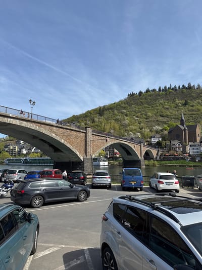 The image depicts a section of Cochem, Germany, showcasing the Skagerrak Bridge, a stone arch bridge spanning the Moselle River. The bridge is partially visible, revealing its arched structure and a pedestrian walkway with railings. On the hill behind the bridge, a church with a tall spire and surrounding buildings are visible. The foreground features a parking area filled with parked cars of various makes, models, and colors. The sky is a clear blue with some light clouds. There are a few people visible on the bridge.