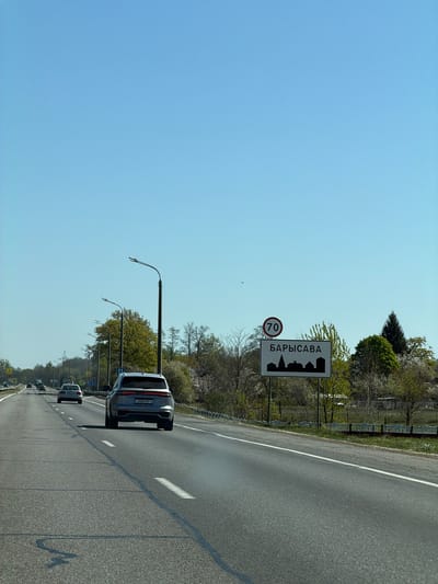 The image depicts a highway in Кіселявецкі сельскі Савет, Belarus, approaching the city of Barysaŭ. A sign indicating the city's name in Cyrillic (БАРЫСАВА) and a speed limit of 70 km/h is visible on the roadside. Cars are traveling on the multi-lane road, with a gray SUV directly in front of the camera. Overhead streetlights are present along the roadway. The sky is blue, suggesting clear weather conditions. Trees and vegetation line the side of the road beyond the sign.