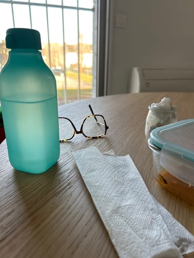 The image depicts an indoor setting, identified as Saint-Étienne, France, featuring a light-colored wooden table surface positioned in front of a window. On the table, from left to right, are a translucent teal-colored plastic water bottle with a darker teal cap, containing water; a pair of folded eyeglasses with tortoise-shell frames; a folded white paper towel with a small bite mark or tear on one end; a small clear glass jar topped with a crumpled white napkin or tissue, possibly containing a condiment; and a clear rectangular plastic food container with a light blue lid, holding an orange-colored food item, with visible condensation inside the lid. The background reveals a large window with vertical mullions, through which golden sunlight illuminates an outdoor urban landscape consisting of blurred buildings, bare trees, and a street. To the right of the window, a plain white wall displays a white wall-mounted air conditioning unit and a light switch. No individuals are present in the scene. The arrangement of items suggests a recent or ongoing meal.