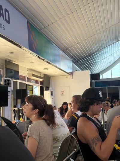 The image shows a crowded waiting area, potentially at an airport or bus terminal, in Del Carmen, Philippines. Multiple people are seated and standing. A man in the foreground wears a black tank top and a backward black baseball cap, showing a tattoo on his upper arm. Behind him, a diverse group of people occupies the available seating. The environment includes a wall with horizontal panels, possibly part of a gate designation, and information displays. Signs indicating "GATE-..." are visible. The image appears to have been taken from a low angle and rotated, making it difficult to ascertain the exact layout of the space.