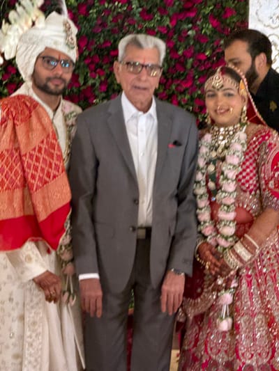 A group of people, including a bride in red and gold traditional attire with floral garlands, and a groom in a white and red patterned outfit with a turban, pose together. An older man in a gray suit and glasses stands in between them. Red flowers are visible in the background. The individuals are positioned in a line, creating a close-knit portrait.