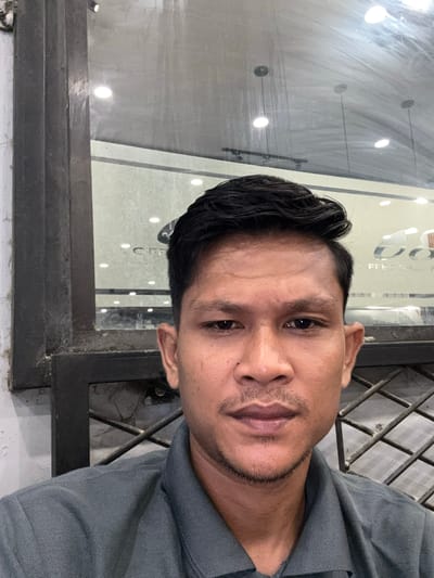 The image displays a chest-up portrait of a South Asian or Southeast Asian man, likely in his late 20s to early 40s, situated in Keudeublang, Indonesia. He is the central subject, looking directly at the camera with a neutral expression, possibly a slight smirk. He has dark, neatly styled hair and light stubble on his chin and upper lip, and is wearing a grey collared polo shirt.
The setting places him in front of a reflective glass panel, suggesting a storefront window or door of a commercial establishment. Visible through and reflected on the glass are several bright, circular ceiling lights, indicating an illuminated interior space. Mirrored text fragments are also reflected on the glass, partially decipherable as "FFEE SU" and "C," hinting at words like "COFFEE" or "OFFICE," reinforcing the commercial context. A robust, dark-colored metal frame or grate is prominent around and behind the man, forming part of the building's structure, possibly a security gate or window frame. The overall impression is an urban environment, with the man captured against a backdrop that suggests an indoor or entryway setting of a shop or cafe. The image itself is rotated 90 degrees clockwise from standard portrait orientation.