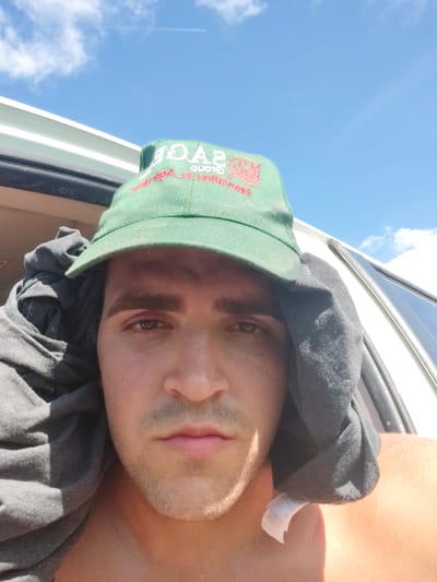 This is a close-up, selfie-style photo of a young man wearing a green baseball cap and a grey t-shirt draped around his head and shoulders. He is outdoors, with a bright blue sky and white clouds visible in the background. A faint contrail from an airplane stretches across the sky. The man is looking directly at the camera with a neutral expression. The green cap has red embroidery on the front, which appears to be text and a logo, possibly related to "Sage" or "Sage Group". A white clothing tag with "XL" printed on it is visible hanging from the t-shirt. The lighting suggests it is daytime, likely midday, due to the bright sunlight and harsh shadows. The overall impression is that of a casual, sunny day. The crops provide closer views of the man's face, the cap's embroidery, and the surrounding sky and vehicle. The location context provided is Magliano Alfieri, Italy.