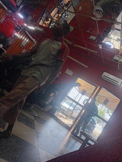 The image depicts an indoor scene in Dorayi, Nigeria, with a partial view of the exterior through a glass entrance. The interior features red painted walls and a geometric, highly reflective ceiling system composed of numerous angled mirror or polished metal panels, reflecting overhead lights and a red sign with "ATTACK" visible. Two white wall-mounted air conditioning units are present on the right wall. The floor is covered with large, alternating light and dark square tiles arranged in a checkered pattern.
In the foreground, a person is captured in motion, appearing blurred. This individual wears a light-colored short-sleeved shirt with red accents on the shoulders/sleeves and light brown trousers, with their back partially towards the viewer. Towards the center-right, a glass door with a dark frame serves as the primary exit/entrance.
Through the glass door, a silhouetted individual is visible standing outdoors near a parked motorcycle. A white vehicle, possibly an SUV, is also visible parked further down the street. The exterior scene is brightly lit by daylight, with trees visible in the background. A yellow and green sign, partially legible as "KARAKI," is affixed to the exterior side of the glass door. A small, dark, round stool or table is positioned inside near the door. The bottom right corner shows a dark, possibly maroon, upholstered object.