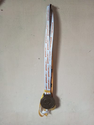 A medal hangs against a textured, light-colored wall. The medal, metallic brown with gold accents, displays a portrait and the word "FINISHER." The medal's ribbon is primarily white, printed with the repeating text "BERLARI," accompanied by "SR." Yellow and gold ribbons are visible beneath the medal. The wall features subtle imperfections and discolorations.