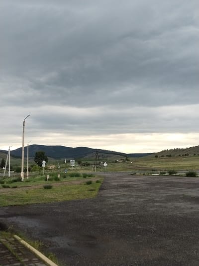 The image depicts a wide, open landscape with a cloudy sky overhead. In the foreground, a dark, wet asphalt surface occupies the lower portion of the frame, with a yellow curb running along the bottom left. To the left of the asphalt, a grassy area with scattered small bushes and posts is visible.
Further into the scene, a road intersects the landscape. Several streetlights stand along the roadside, with one prominent, tall pole on the left. Various road signs are present, including a triangular warning sign with a number "60" below it on the left, and a circular sign further back.
The middle ground features rolling green hills dotted with trees and some scattered buildings. The background consists of larger, darker, forested hills that stretch across the horizon. The overall impression is a somewhat overcast, rural environment, possibly on the outskirts of a settlement.
The location is identified as the city of Moskovskoye, Russia.