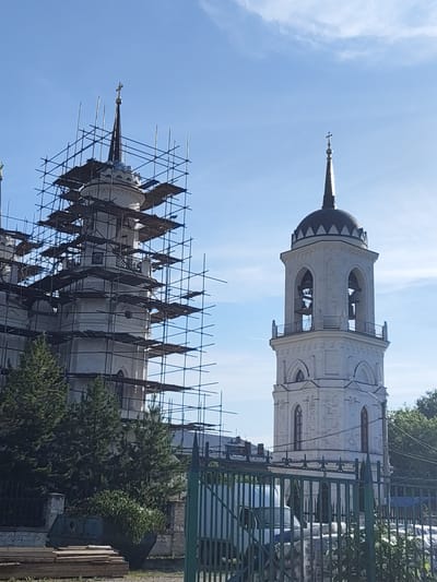The image displays two white stone towers of the Vladimirskaya Church in Bykovo, Moscow Oblast, Russia, against a clear blue sky with minimal clouds. The tower on the left is extensively covered in dark metal scaffolding, indicating ongoing construction or renovation; its dark spire is topped with a golden cross. The fully visible tower on the right, a bell tower, features multiple levels with arched openings revealing bells, capped by a dark, segmented dome and a dark spire with a lighter-colored cross. In the immediate foreground, a dark green metal fence is visible, partially obscuring a white vehicle. Evergreen trees are situated between the fence and the scaffolded tower, with a large green industrial dumpster and stacked wooden planks visible at the base of the construction area. No people are present.