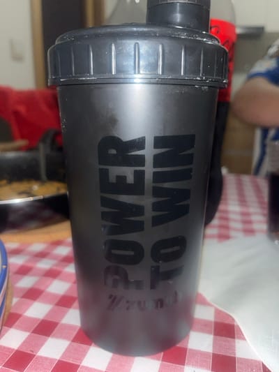 The image shows a black cylindrical shaker bottle prominently placed on a red and white checkered tablecloth within an indoor setting in the city of Pedrouços, Portugal. The shaker bottle features the vertically oriented text "POWER TO WIN" in black capital letters, with a faint "Zumub" logo visible below. Its black screw-on lid has a smaller, black pop-top closure, and both the lid and upper body exhibit light white residue. In the blurred background to the left, a dark cooking pan with food and a red textile are partially visible. To the right, a red beverage bottle and a glass with a dark liquid are present, along with the upper arm of an individual wearing a blue and white striped shirt. A small segment of a blue and white patterned dish is visible in the lower left foreground.