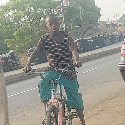 A young male, appearing pre-adolescent, is positioned on a bicycle on a paved road in Gudi, Nigeria. He wears a brown and white striped short-sleeved shirt and loose-fitting teal shorts, with a short haircut. His mouth is slightly open, and he looks towards the right side of the frame, hands gripping the bicycle handlebars. The bicycle, an older model, appears to have a red or pink frame with parts of the front wrapped in light-colored material, and is equipped with knobby tires.
To the male's left, beyond a concrete curb bordering the road, multiple large, dark-colored sacks are stacked along the roadside, suggesting contents like charcoal. Green trees are visible in the background. To the male's right, the rear section of a dark-colored vehicle with red taillights is partially visible. Further back, a low concrete barrier runs parallel to the road, behind which more stacks of dark sacks are present. Distant elevated structures or bridge supports are discernible under bright daylight conditions.