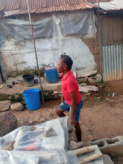 In Karsana 2, Nigeria, a young girl with braided hair, wearing a red polo shirt and denim shorts, stands on a dirt path next to a wall. The wall appears to be made of concrete and mud, with sections of exposed brick. Above the wall is a corrugated metal roof with a rustic, rusted appearance. The girl is looking to her right, with her left leg bent and her right leg slightly forward.
In the background, to the left of the girl, are several objects: a black bucket, a small purple container, and a large blue plastic barrel containing some pink and patterned fabric. A broom and a pair of black sandals are placed on a step behind the black bucket. To the right of the girl, a large blue plastic bucket is positioned on the ground.
In the foreground, a flat surface is covered with clear plastic sheeting, on which are placed several rocks and pieces of bamboo. The ground is a mixture of dirt and small stones. The overall environment suggests an outdoor setting in a potentially informal or developing urban area, characterized by weathered structures and everyday objects.