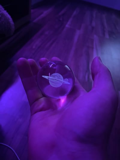 The media file shows a hand holding a clear glass orb that contains a white etched image of a planet with rings. The orb is illuminated by purple light, which casts a glow on the hand and the background. The background appears to be a wooden floor, with some shadows and lighter areas, also illuminated by the purple light. There is a hint of a dark object in the lower left corner of the image and some white string or cable in the lower right corner. The overall mood is somewhat mysterious and ethereal due to the lighting. The specific planet depicted is not definitively identifiable but resembles Saturn. The setting is indoors, with the purple lighting suggesting it could be evening or a dimly lit room. No text is visible in the image.